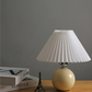 Beige table lamp with pleated white shade on a wooden surface against a gray wall