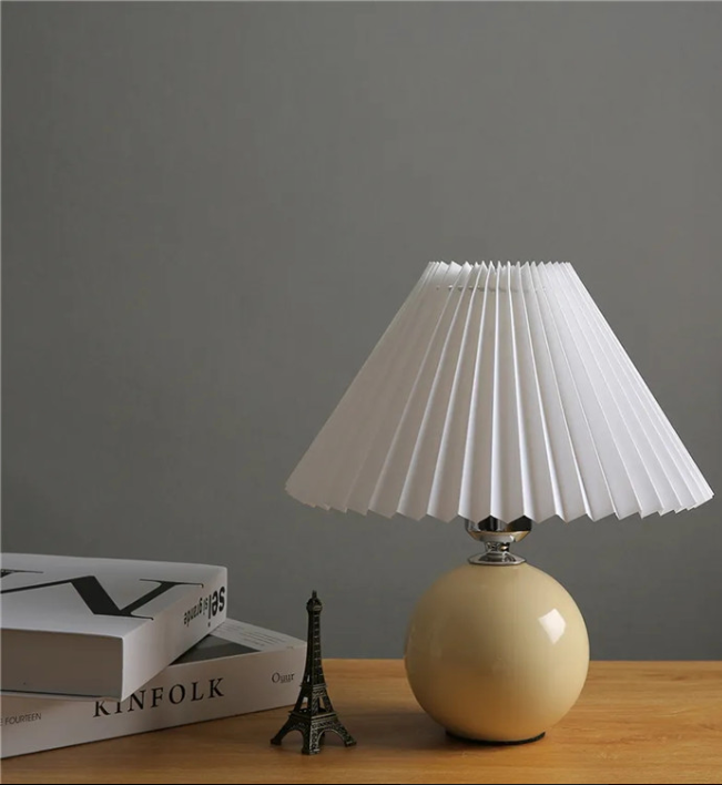 Beige table lamp with pleated white shade on a wooden surface against a gray wall