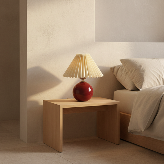 Red lamp with a beige shade on a wooden nightstand next to a bed.